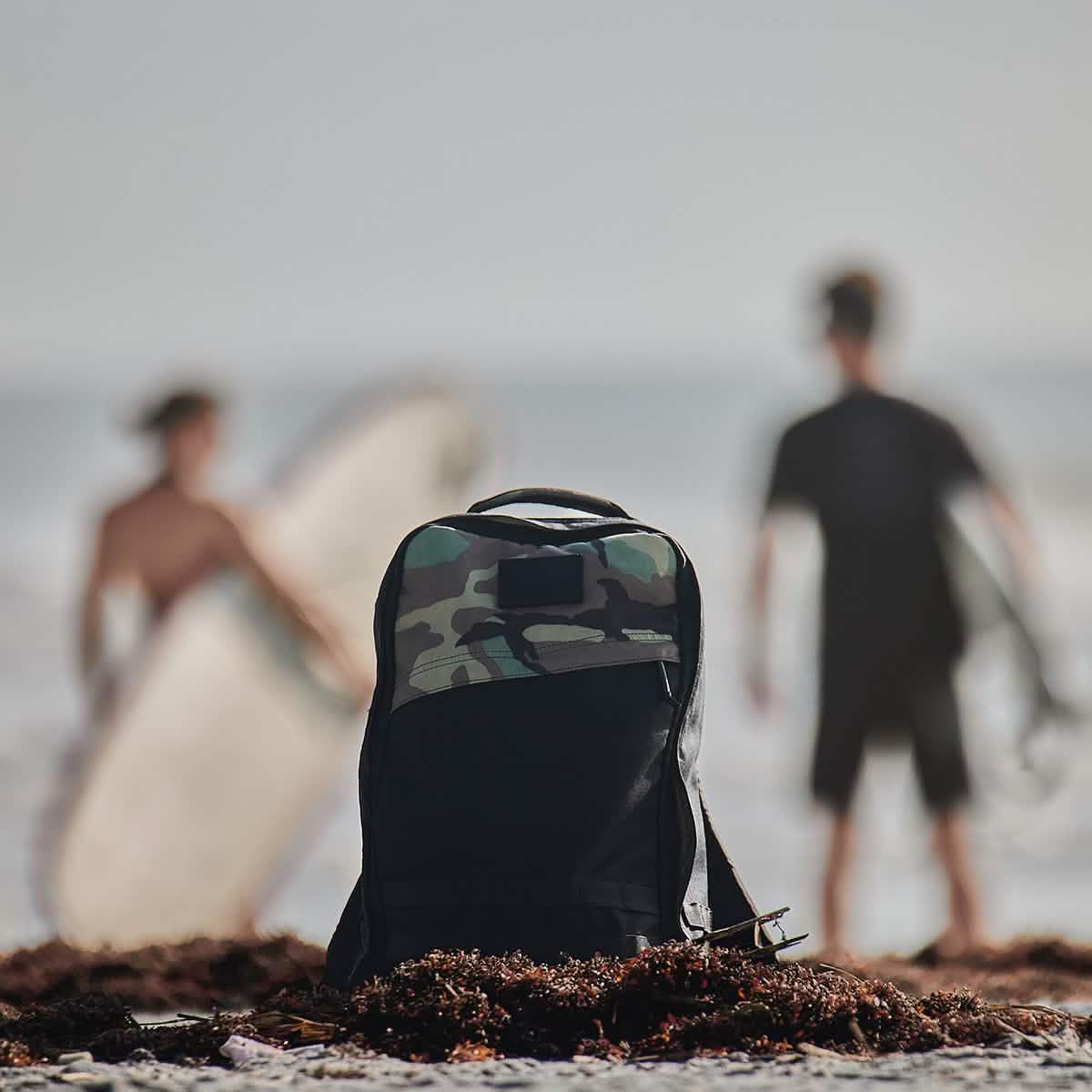 Black and camo GORUCK backpack on sandy beach with two surfers holding surfboards in background
