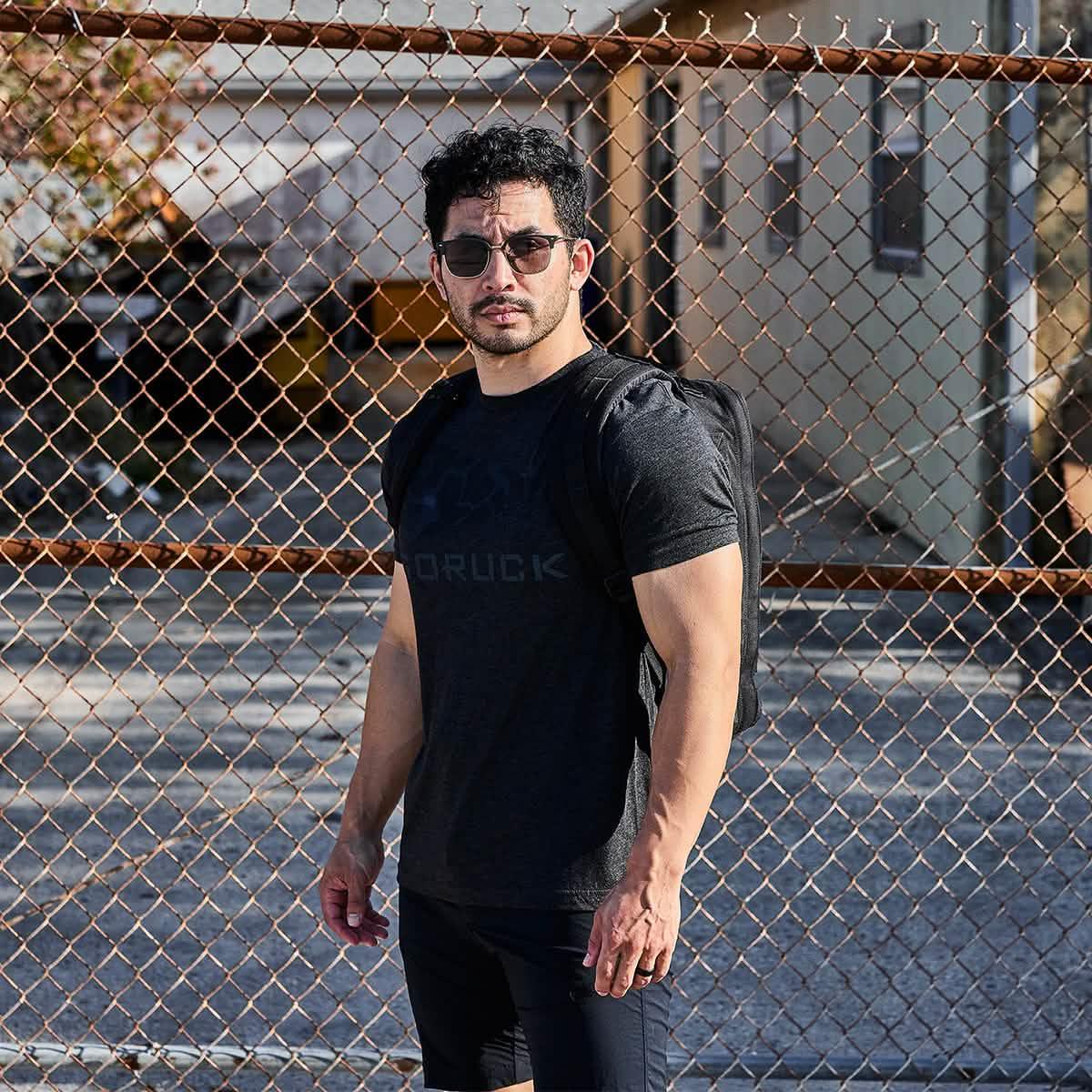 Man wearing black GORUCK t-shirt and backpack standing in front of metal chain-link fence in urban setting