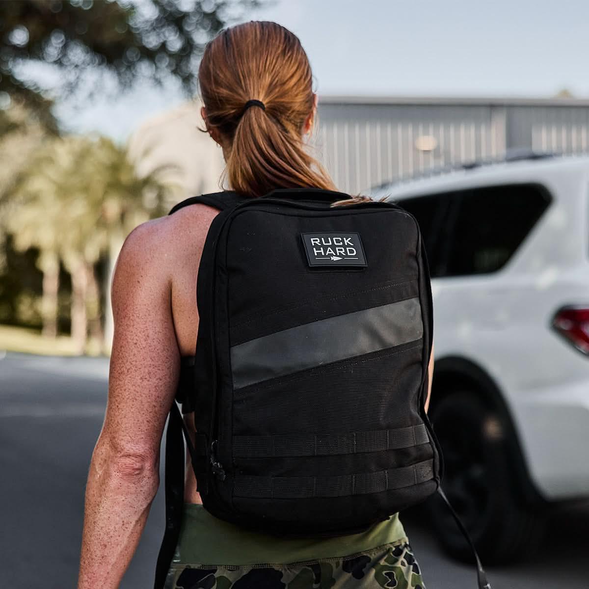 Person with red hair in a ponytail wearing a black rucksack labeled 'Ruck Hard' outdoors near a white vehicle