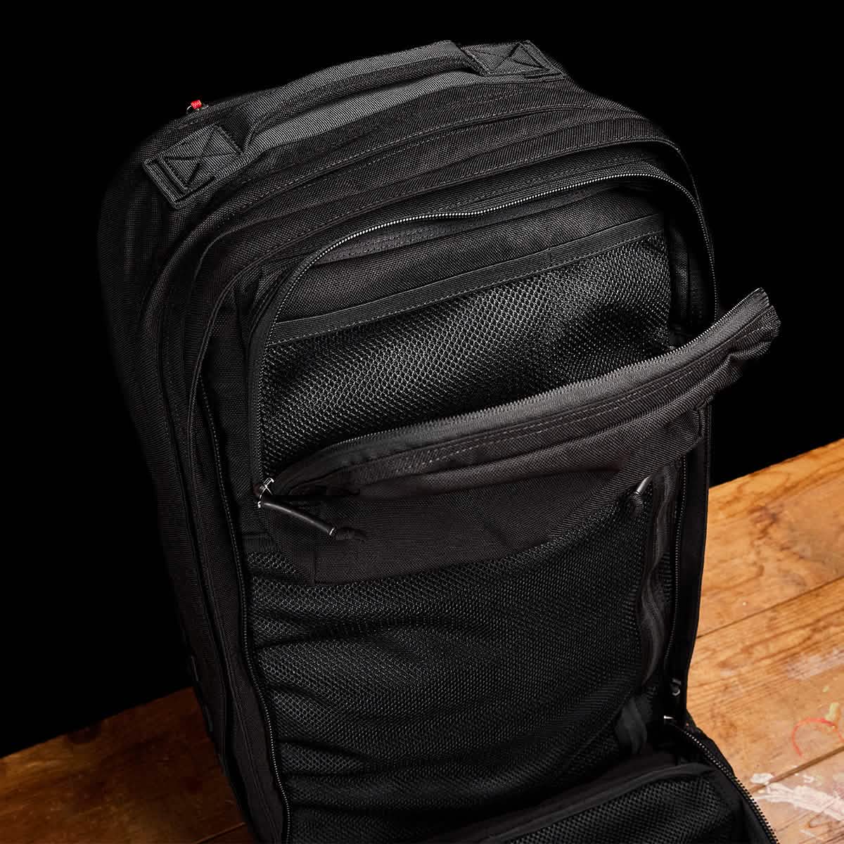 The GR2 travel bag by GORUCK is open on a wooden surface, revealing several compartments and zippers. It seems to be crafted from durable fabric with mesh sections for organization. The background is a plain black.