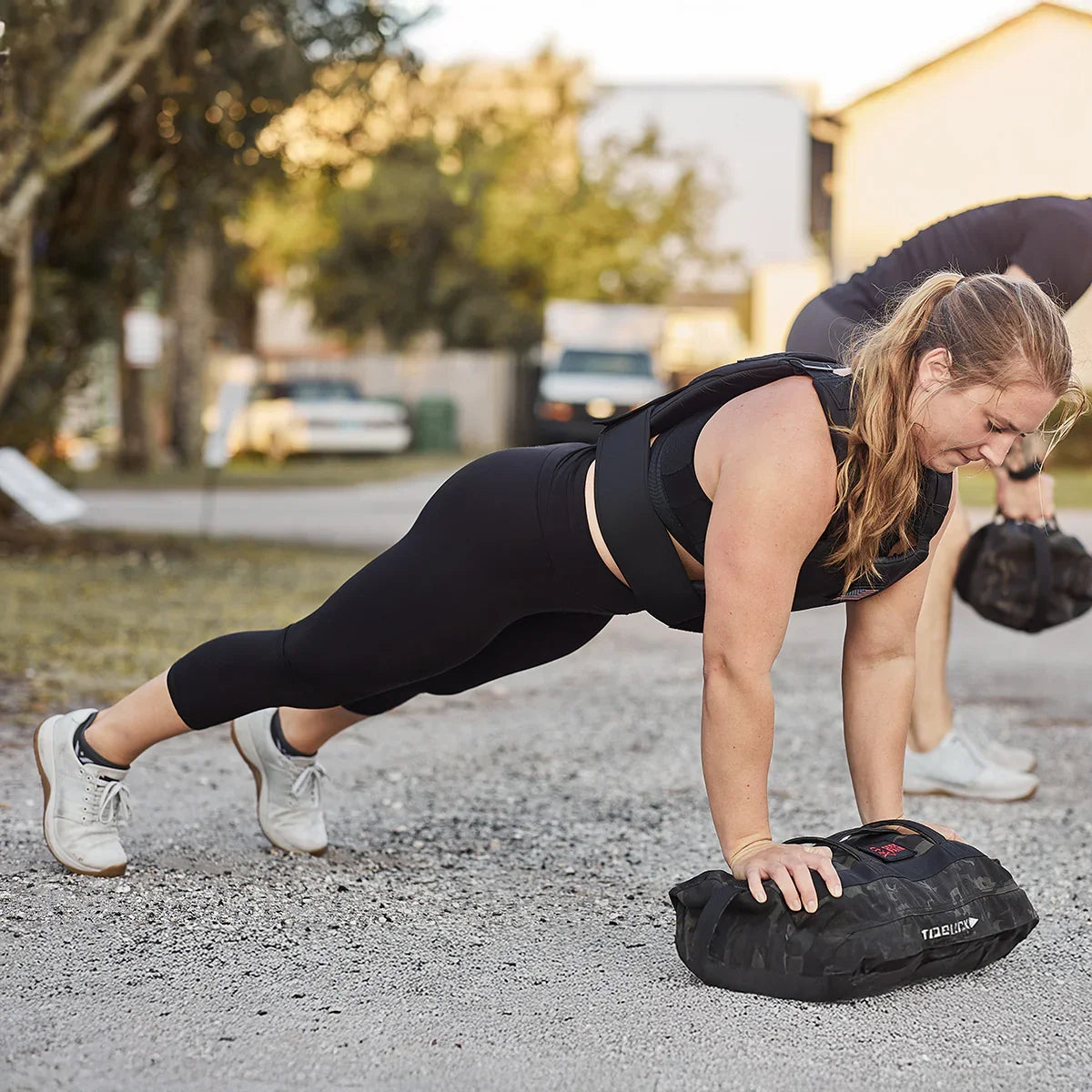 Woman in black leggings and workout vest doing plank exercise outdoors using a weighted rucksack on gravel