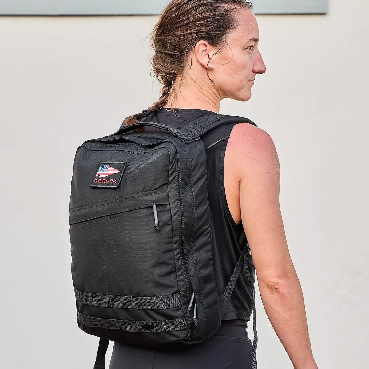 A woman with braided hair wears a GR0 - Ripstop ROBIC® compact rucksack and a sleeveless top, standing against a light background.