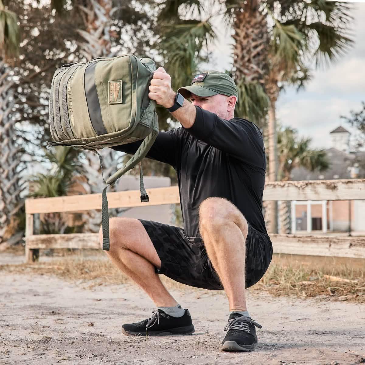 Man in athletic gear doing a squat while holding a green tactical GORUCK backpack outdoors near palm trees