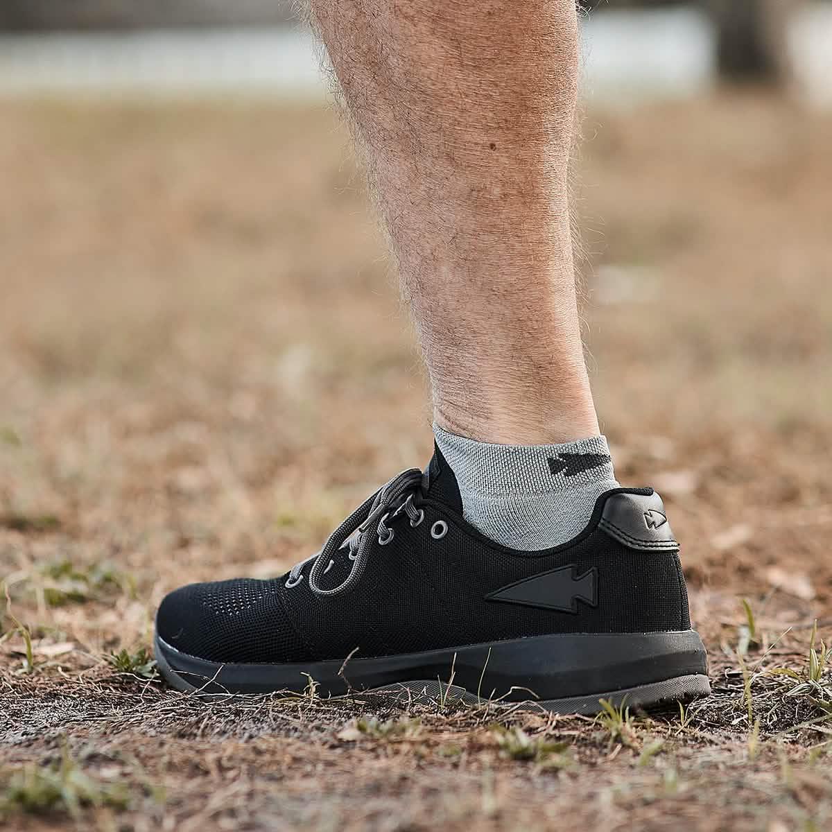 Side view of a person wearing black GORUCK rucking shoes and gray ankle socks outdoors on dry grass