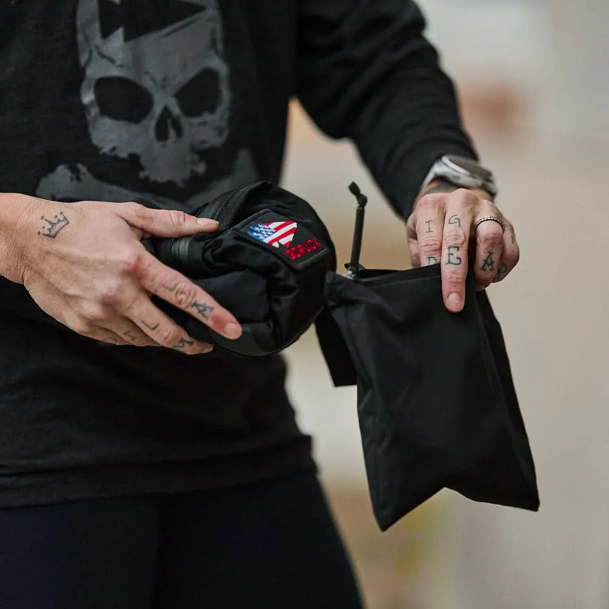 A person with tattooed hands holds a black Packable Bullet Ruck - Ripstop ROBIC® and a folded item featuring a USA flag patch.