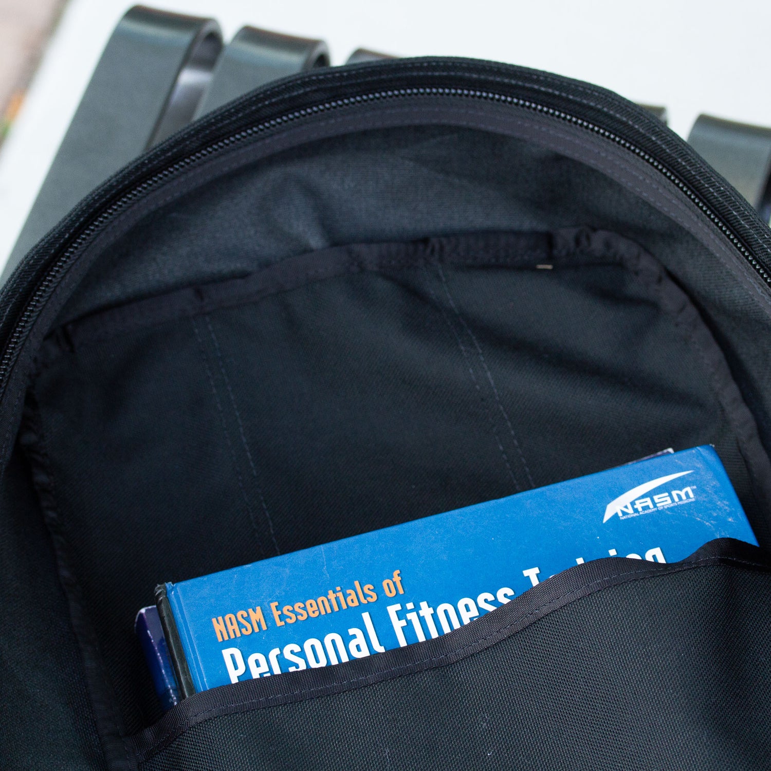 A blue fitness training book peeks out from the front pocket of an open black Shadow Ruck backpack, designed for everyday durability and carry.