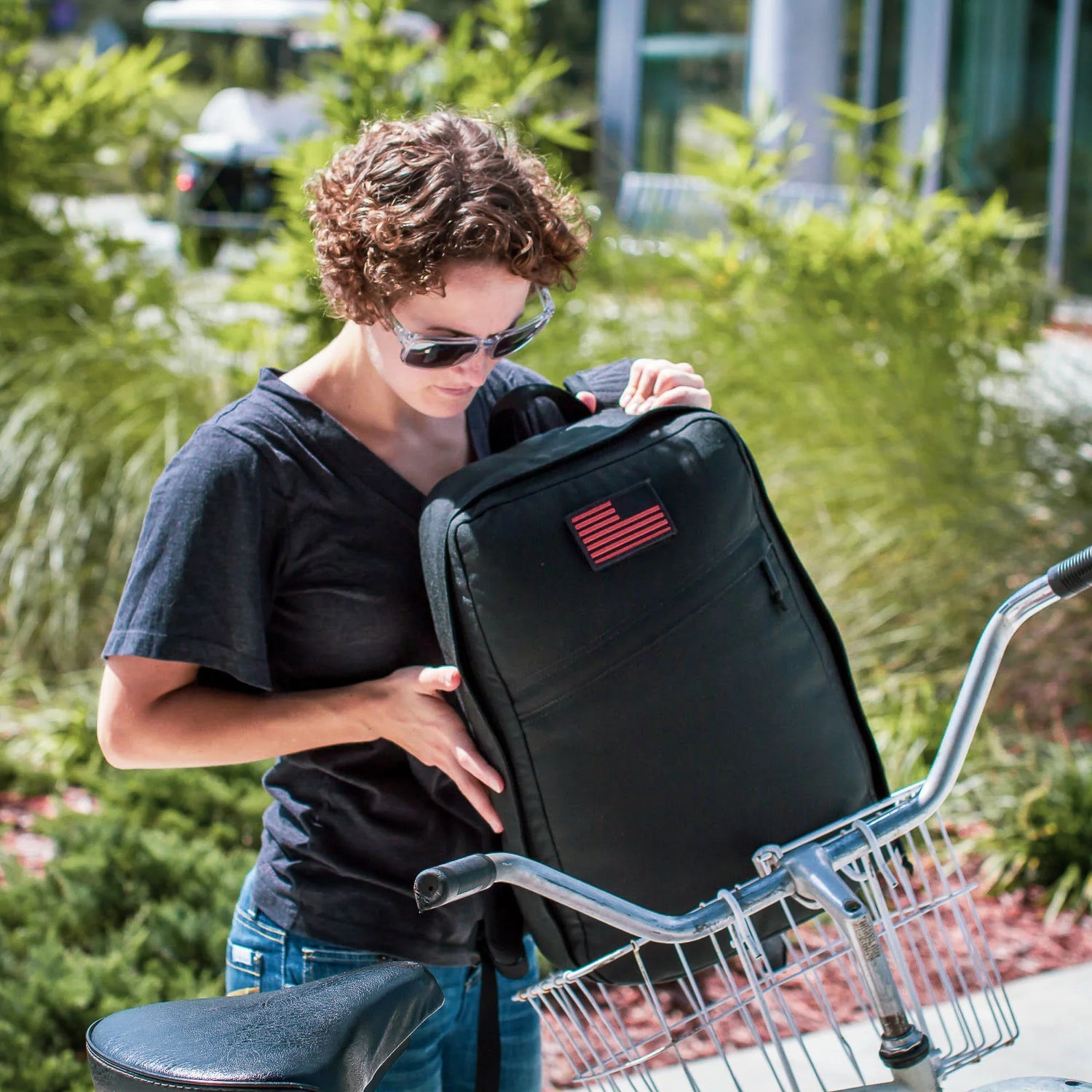 Woman placing GORUCK black rucker backpack with USA flag patch into bicycle basket outdoors