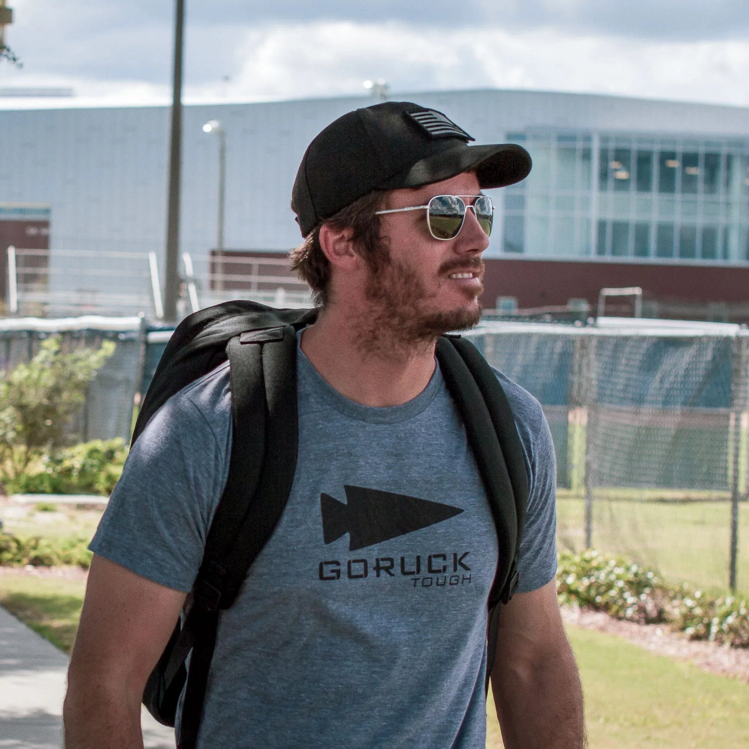 Man wearing GORUCK Tough shirt, cap, and backpack outdoors in athletic setting