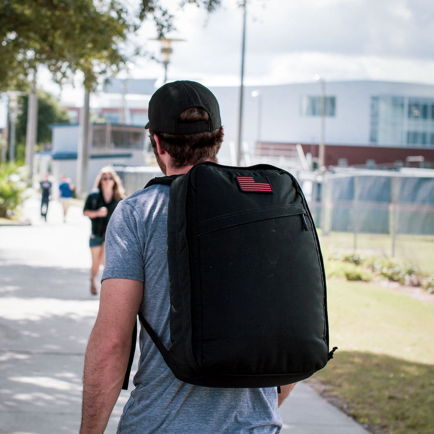 A man walks outdoors on a sunny day, carrying the Shadow Ruck—a lightweight backpack featuring a flag patch.