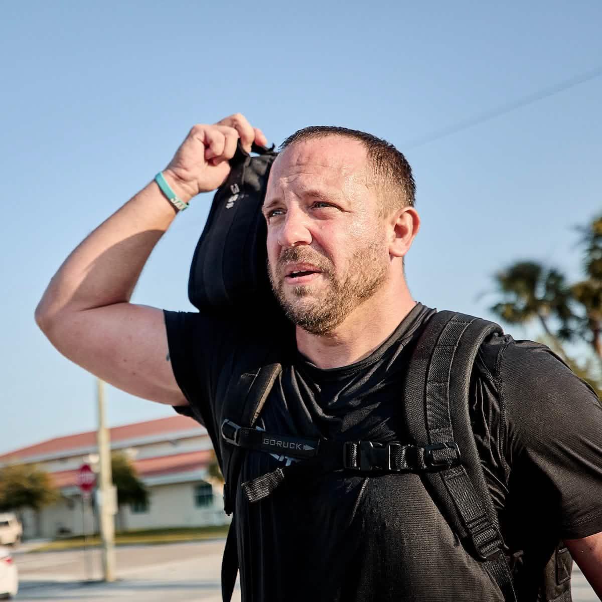 Man wearing black GORUCK Tough Mesh Tee and rucksack, wiping sweat outdoors under clear sky