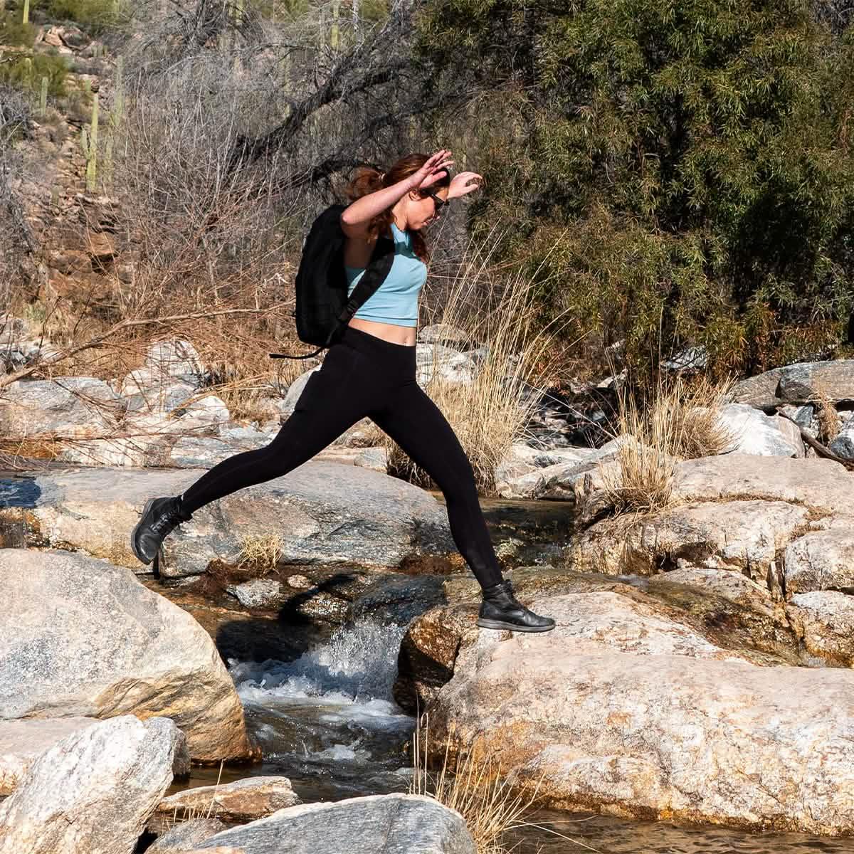 Woman wearing GORUCK backpack jumping across rocks over a stream in a dry, rocky outdoor setting