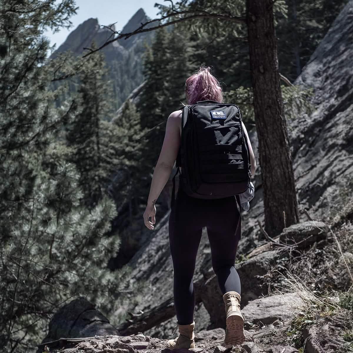 Hiker with GORUCK black tactical backpack walking on rocky forest trail with mountains in background