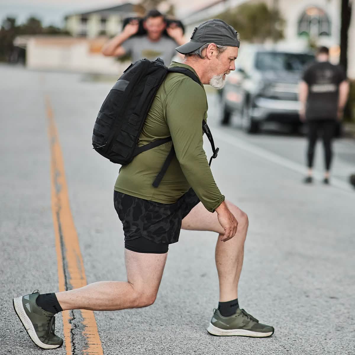A man wearing a GORUCK Rucker 4.0 performs lunges on a street. He is clad in a green long-sleeve shirt, black shorts, and a cap worn backward. In the background, another individual walks down the road with a child on their shoulders.