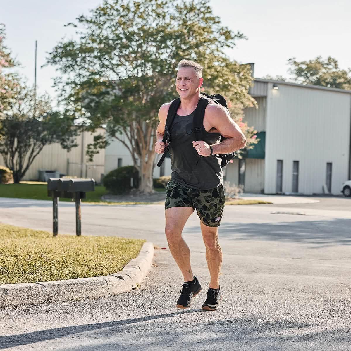 Fit man running outdoors wearing camouflage shorts, black tank top, and a rucking backpack on a sunny day