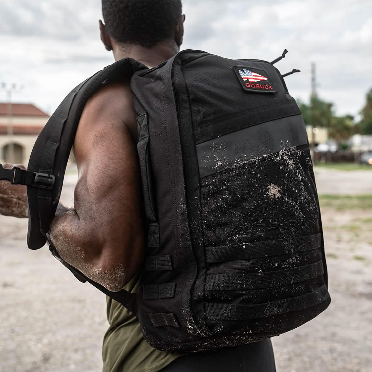 Muscular man wearing a muddy black GORUCK backpack outdoors with a cloudy sky background