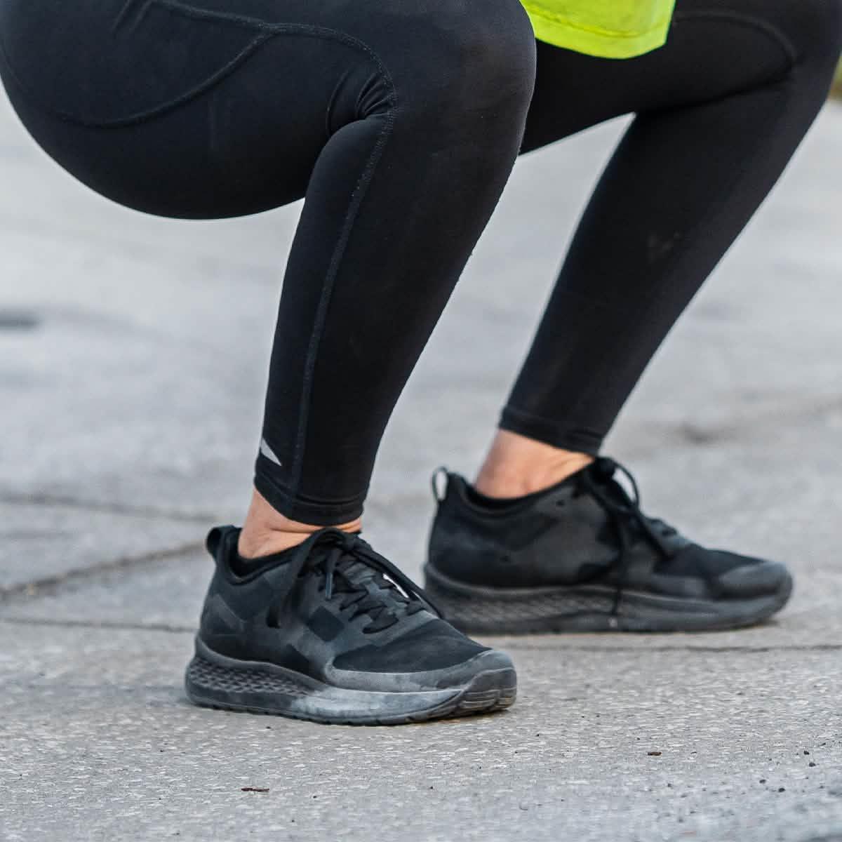 Person wearing black athletic leggings and black sneakers squatting on concrete surface