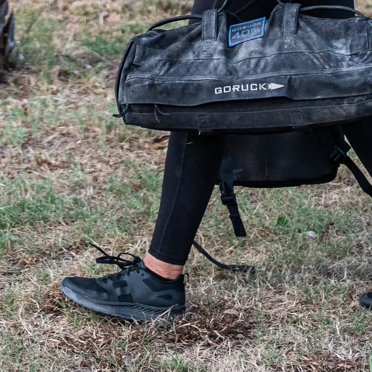 Clad in the Women's Rough Runner - Blackout black leggings and sneakers featuring an EVA midsole, a person carries a large black GORUCK bag over their shoulder while walking on grass. The bag displays visible GORUCK branding and boasts a rough, rugged look ideal for outdoor activities.