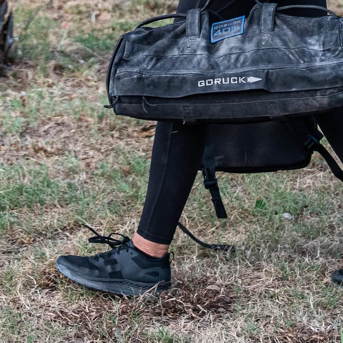 Person wearing black athletic shoes and leggings carrying a black GORUCK duffel bag outdoors on grass