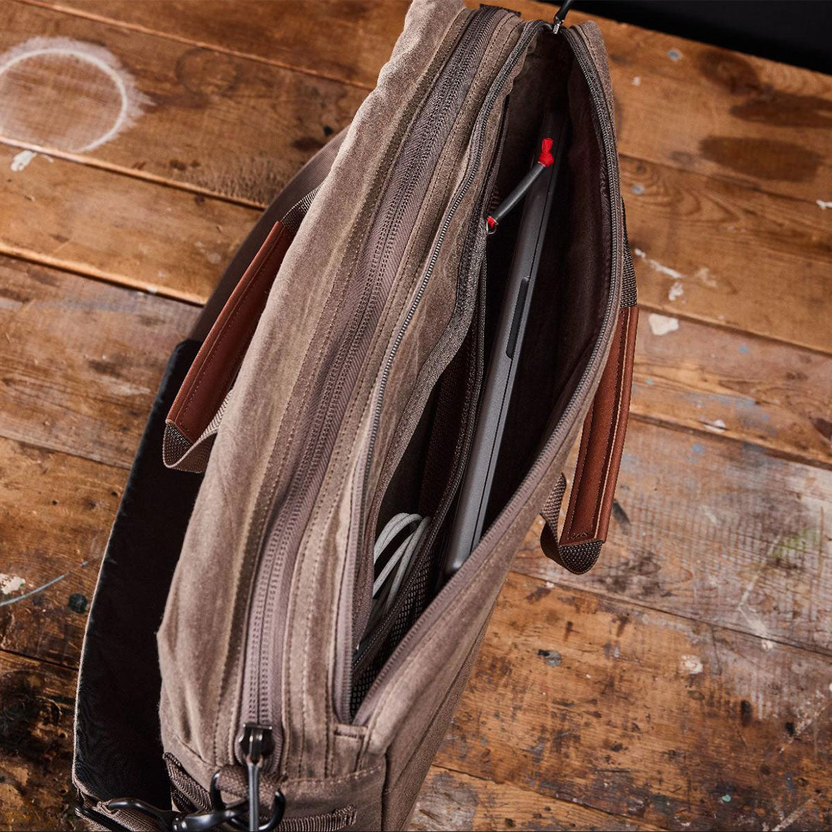 Top view of an open brown messenger bag on a wooden surface, showing a laptop and cables inside.