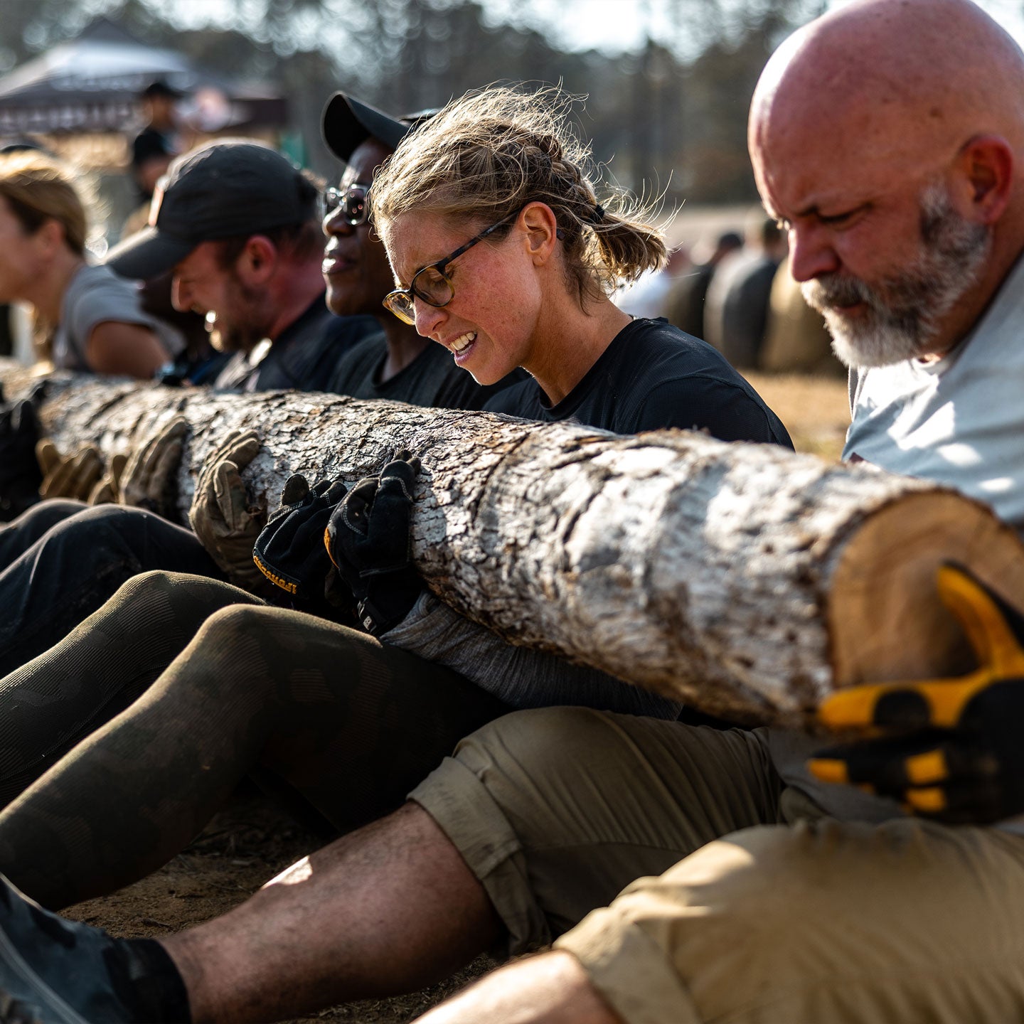 A group of people work together, lifting a large log outdoors during a team fitness challenge.