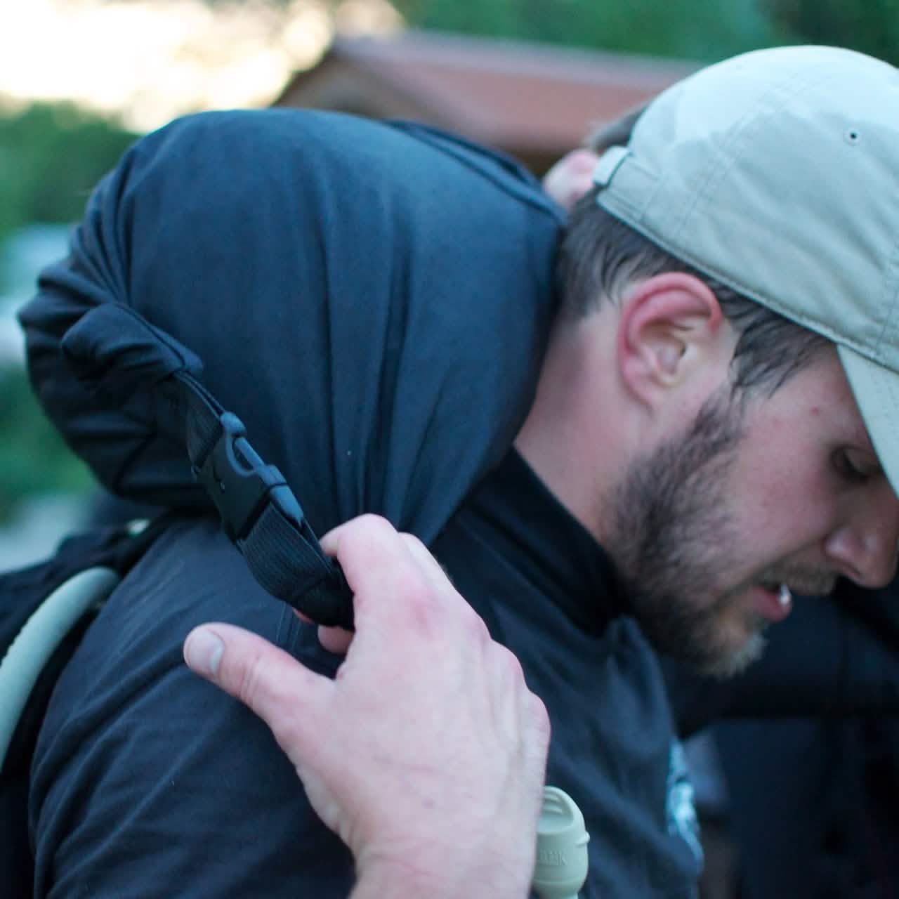 A man dressed in a black shirt and beige baseball cap carries a GORUCK Brick Bag, with its stuff sack wrapped around the shoulder strap. He seems to be outdoors, and his face is partially turned away from the camera.