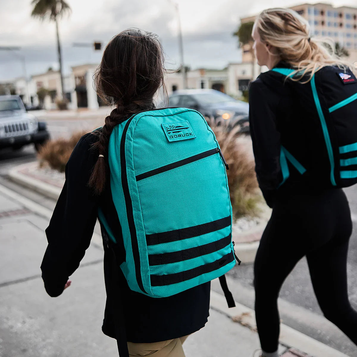 Two women walking on city sidewalk wearing black outfits with teal and black GORUCK backpacks