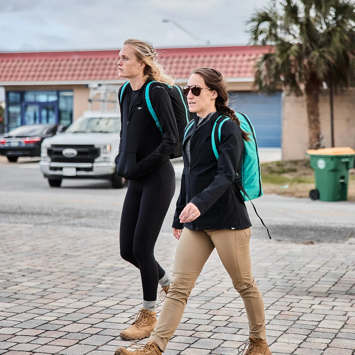 Two women walking outdoors wearing black jackets, tan boots, and teal GORUCK backpacks on a paved street