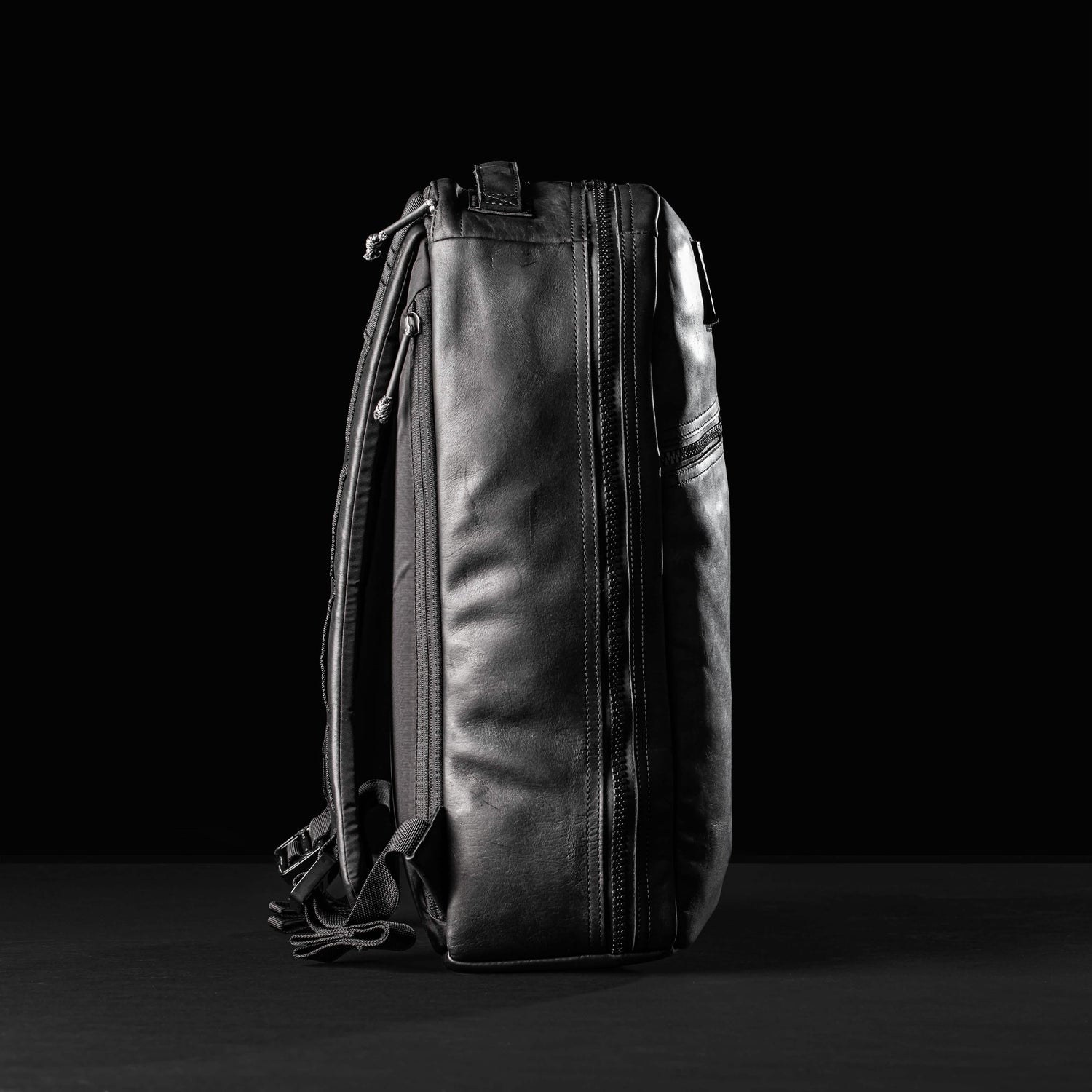 Side profile of a black leather GORUCK Bullet 15L backpack with zippers and shoulder straps on black background