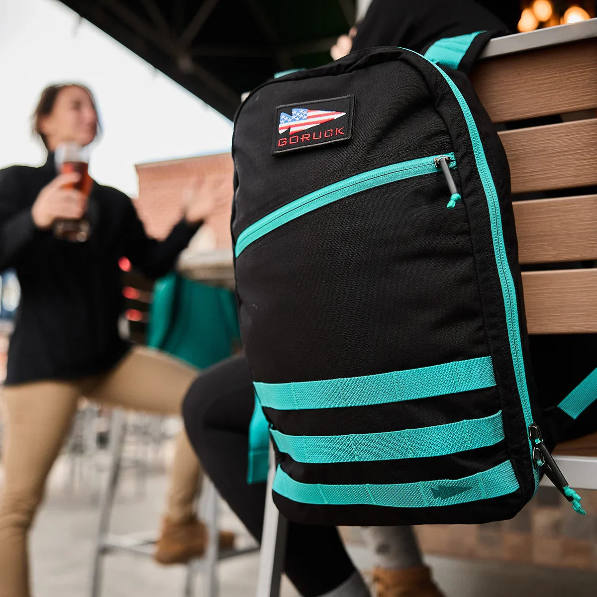 Black GORUCK Bullet 15L backpack with teal accents hanging on a chair outdoors, blurred person holding a drink in background