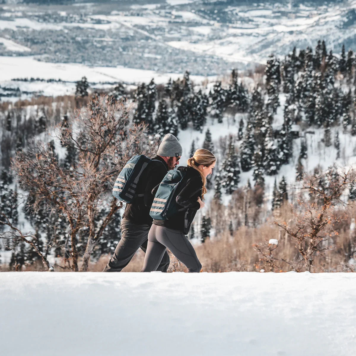 Two people rucking with GORUCK backpacks on a snowy trail surrounded by winter trees and hills