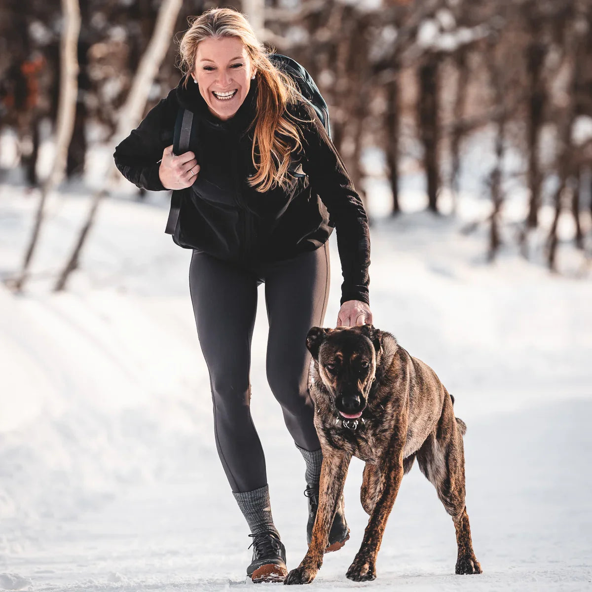 Smiling woman in black activewear with backpack walking a large brindle dog on snowy trail