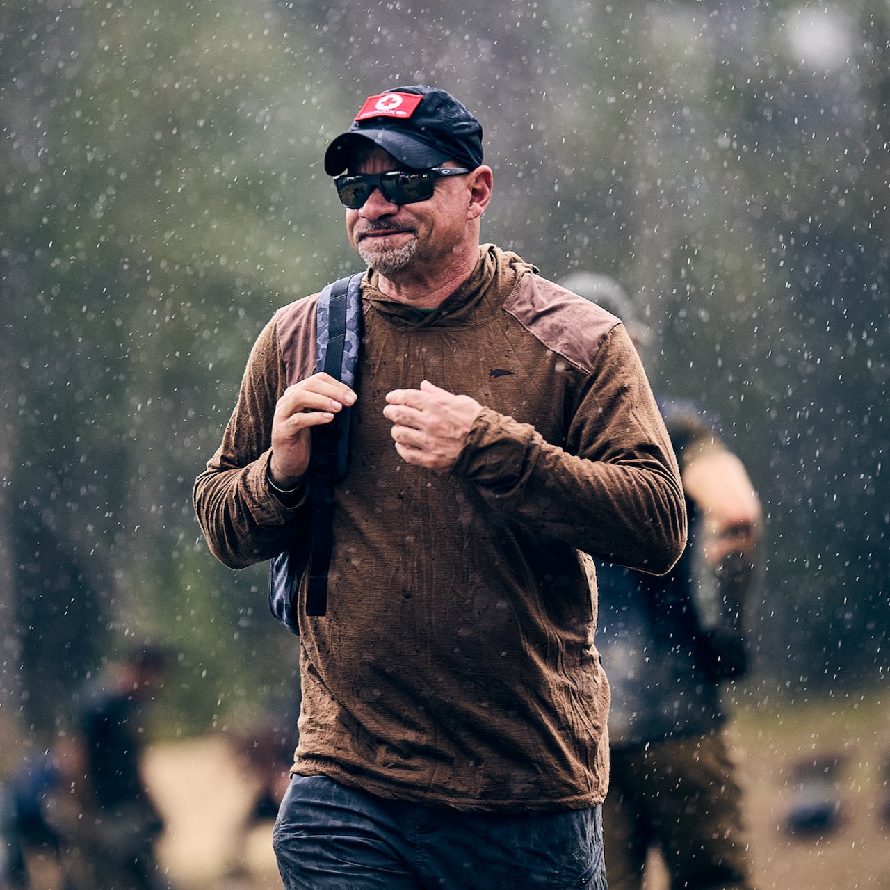 Man wearing a cap and sunglasses walks in the rain with the Double Compartment Bullet Ruck - Ballistic Nylon Cordura - 18L, featuring a bombproof laptop compartment; blurred people appear in the background.