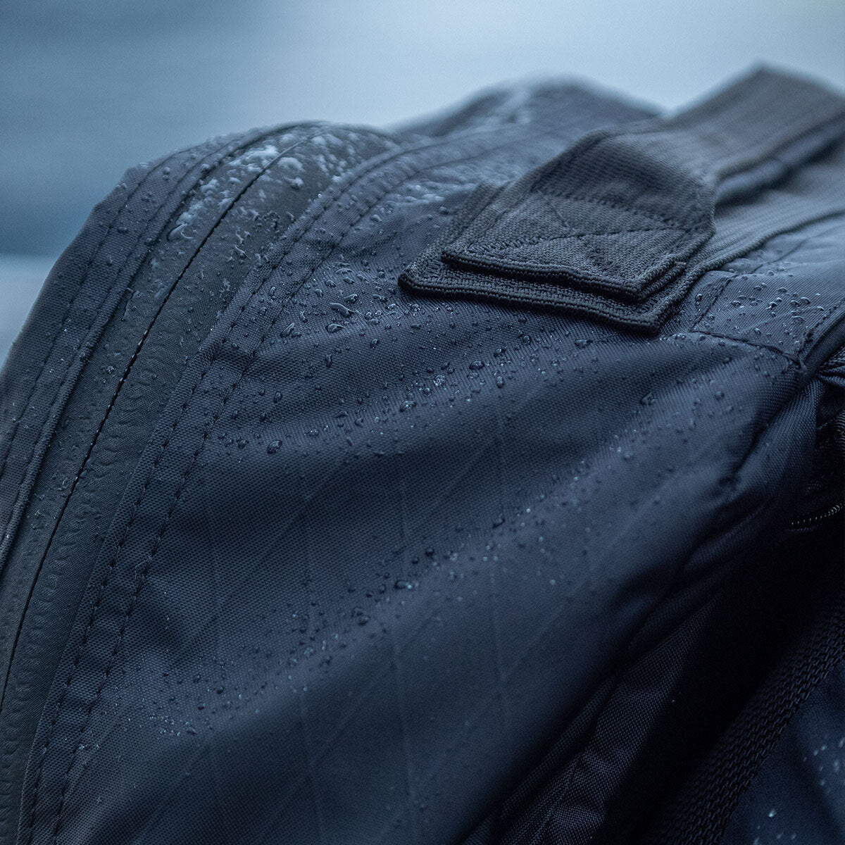 Close-up of a black fabric backpack with water droplets on its surface, suggesting it is water-resistant.