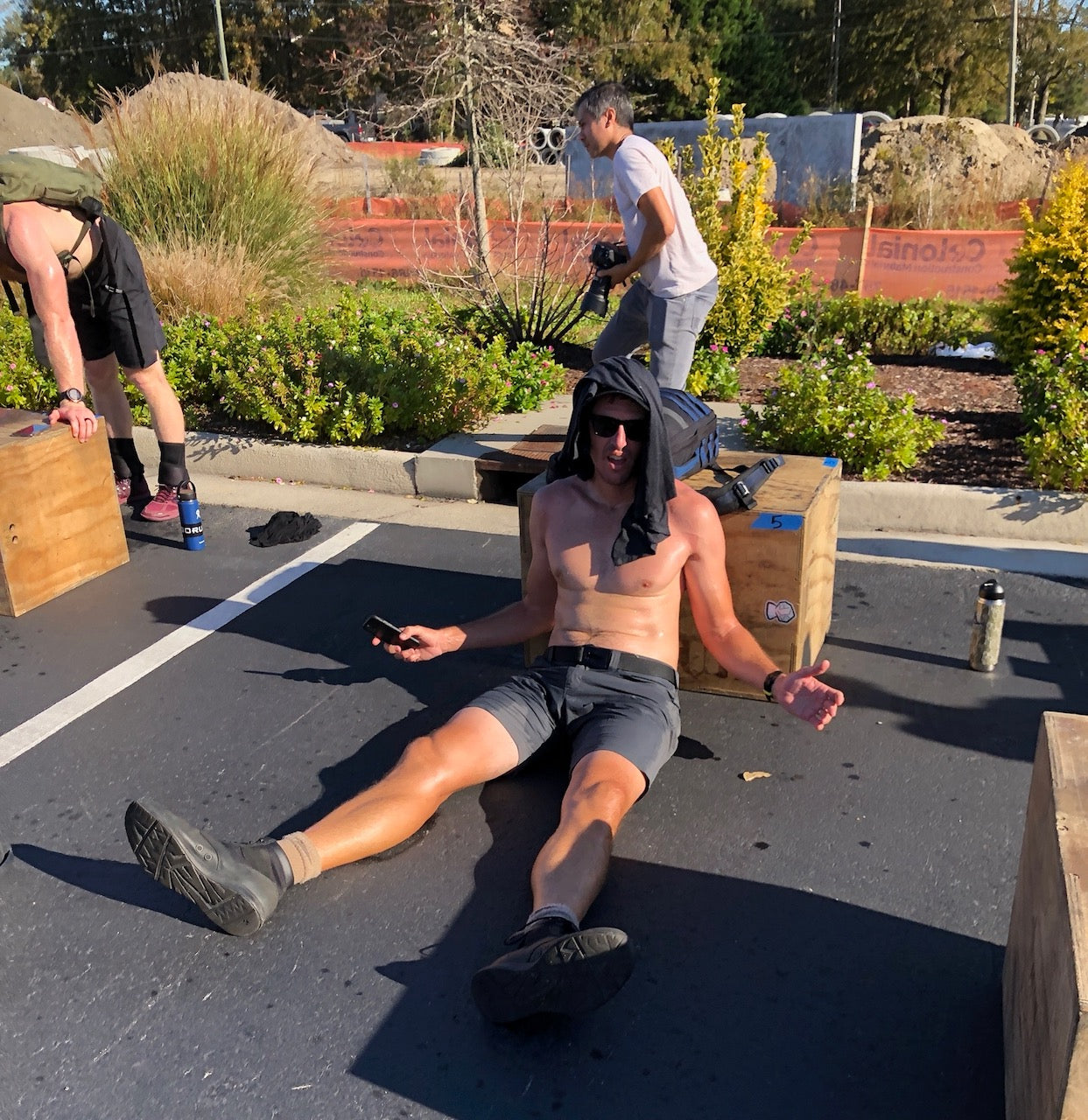 A man wearing Men’s Simple Shorts - Lightweight ToughDry® sits on the pavement, resting after his workout with a shirt over his head while others gather nearby.