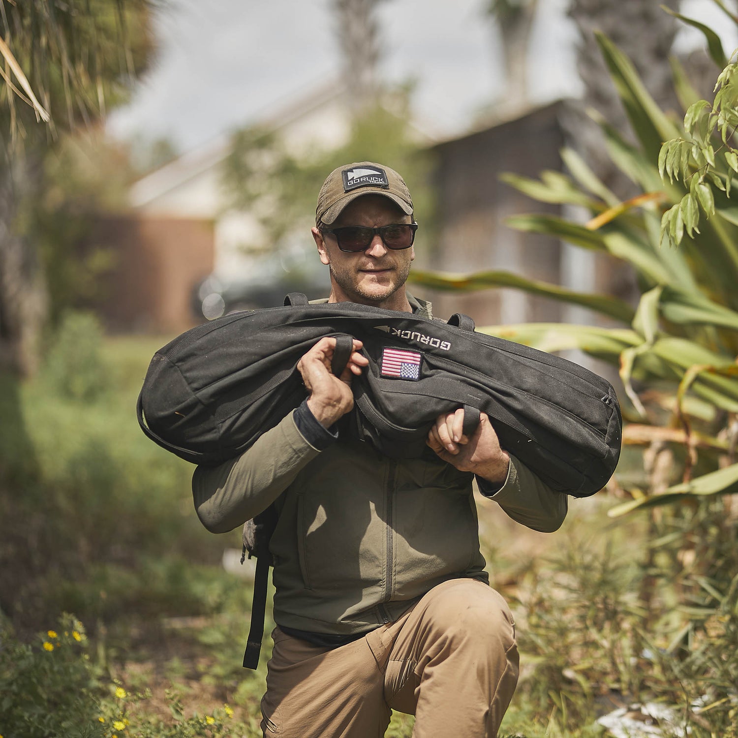 A man in sunglasses and a cap carries a large black duffel bag outdoors in a garden, wearing the abrasion-resistant Indestructible Challenge Windbreaker.