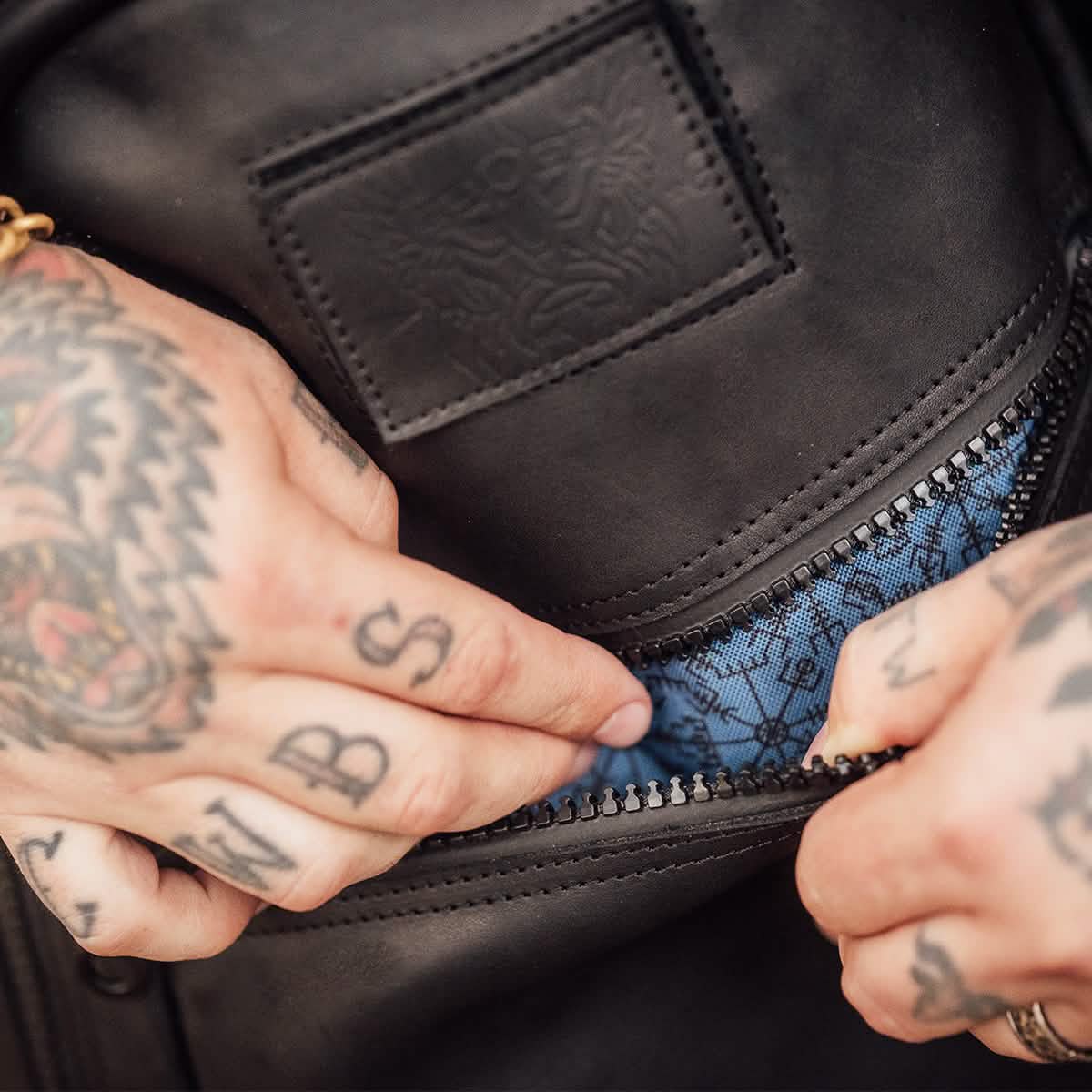 Close-up of tattooed hands unzipping the GORUCK Bullet Ruck x Carryology - Berserker Viking, unveiling its blue patterned interior lining. The visible tattoos featuring an array of letters and designs on the fingers suggest a Berserker Viking theme.