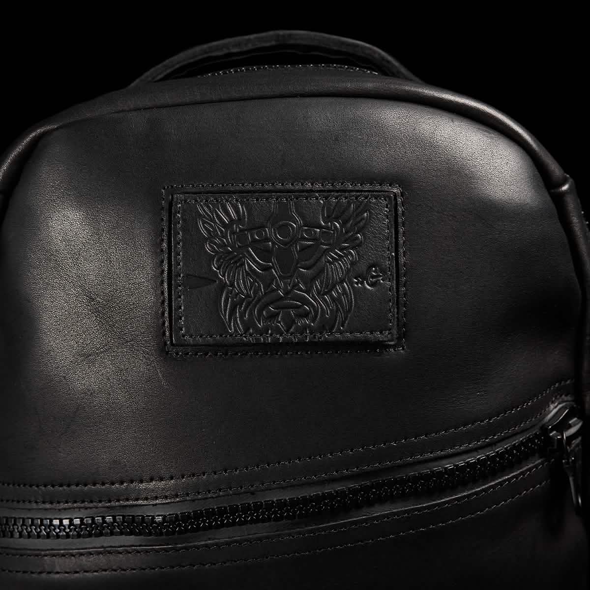Close-up of a GORUCK Bullet Ruck x Carryology - Berserker Viking black leather backpack with an embossed patch showcasing a detailed design. The patch features intricate motifs that resemble a stylized Berserker Viking face, with a zipper below it against the dark background, highlighting its robust construction.