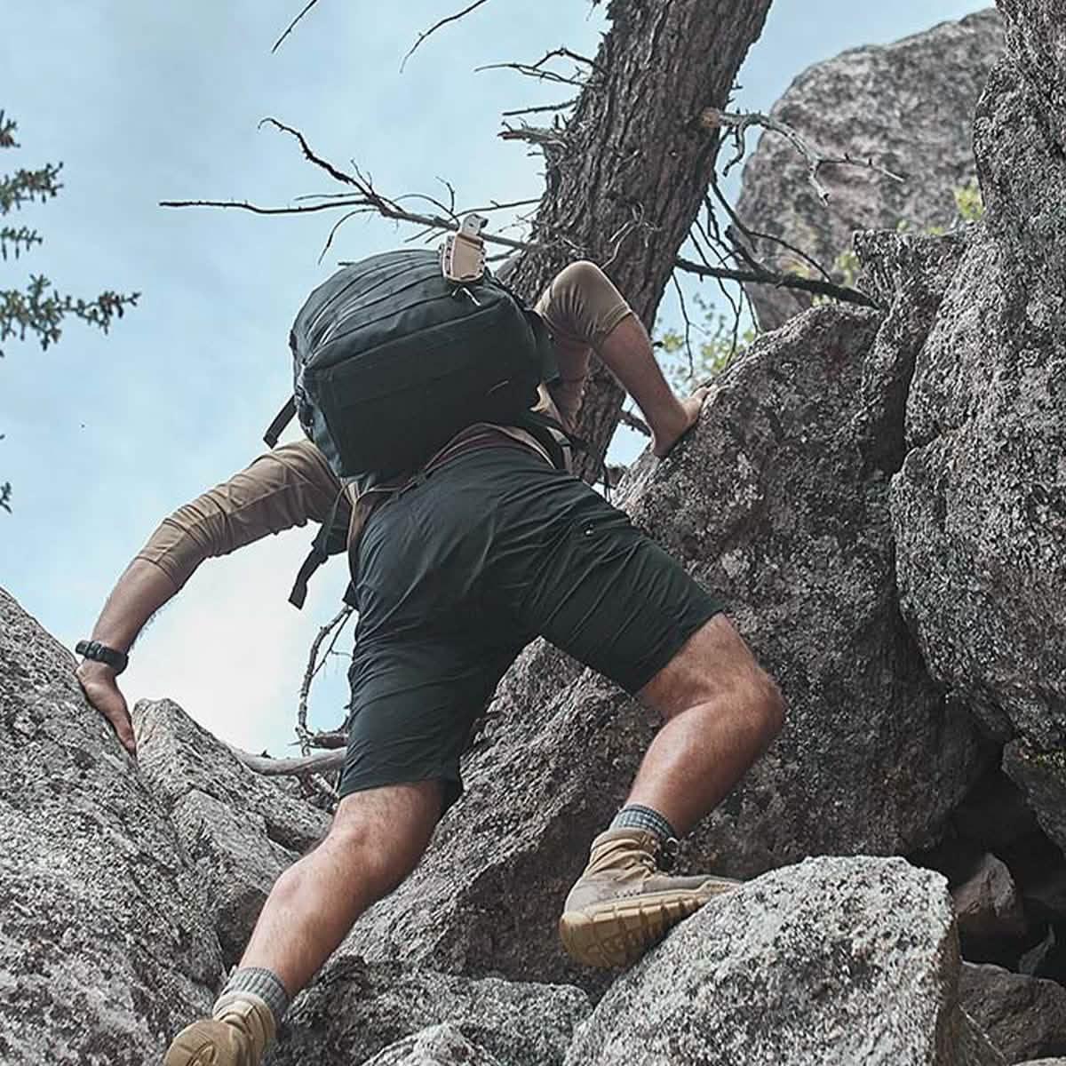 Person rock climbing outdoors wearing black GORUCK shorts, tan hiking boots, and carrying a large backpack