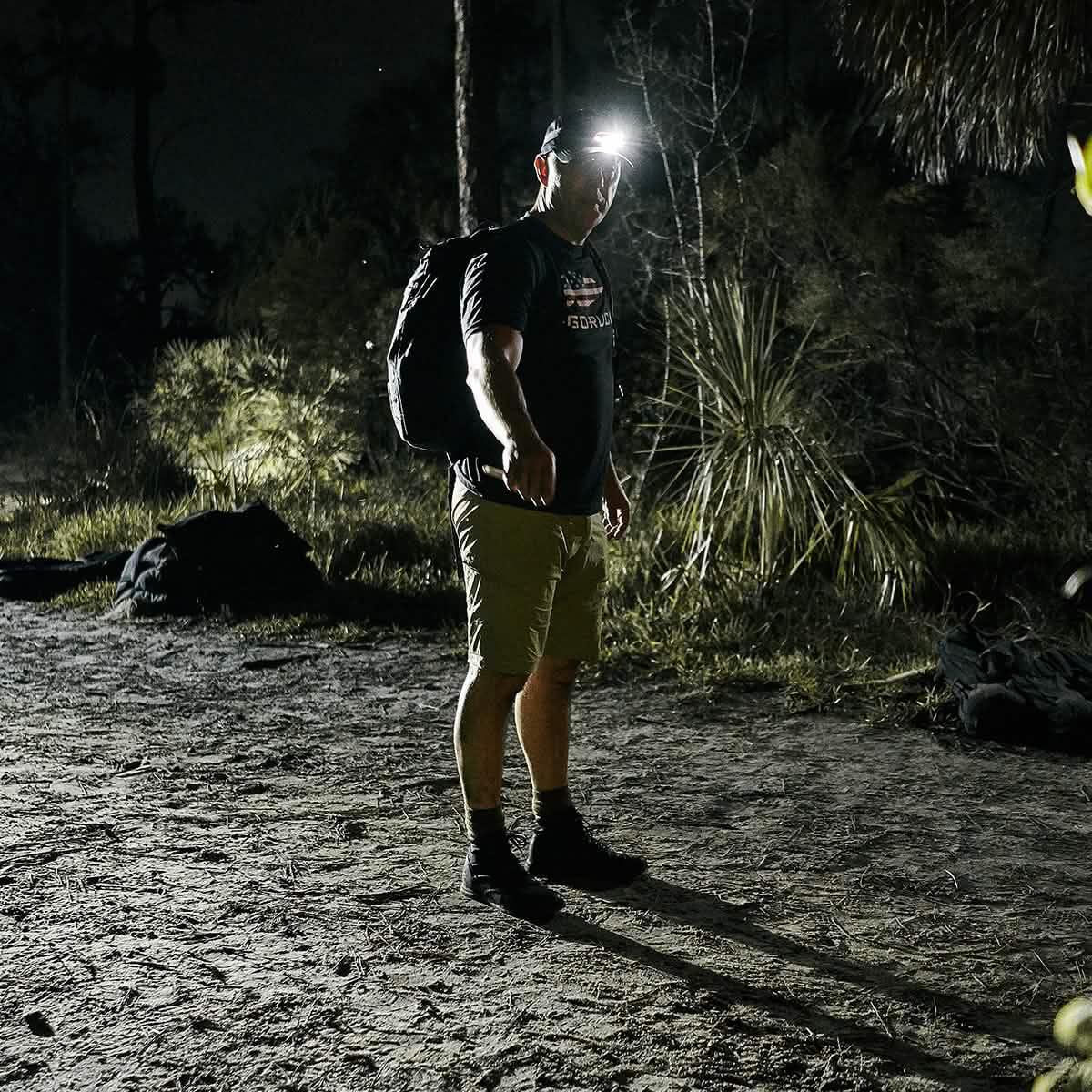Man wearing GORUCK gear with backpack and headlamp standing on dirt trail at night in forest