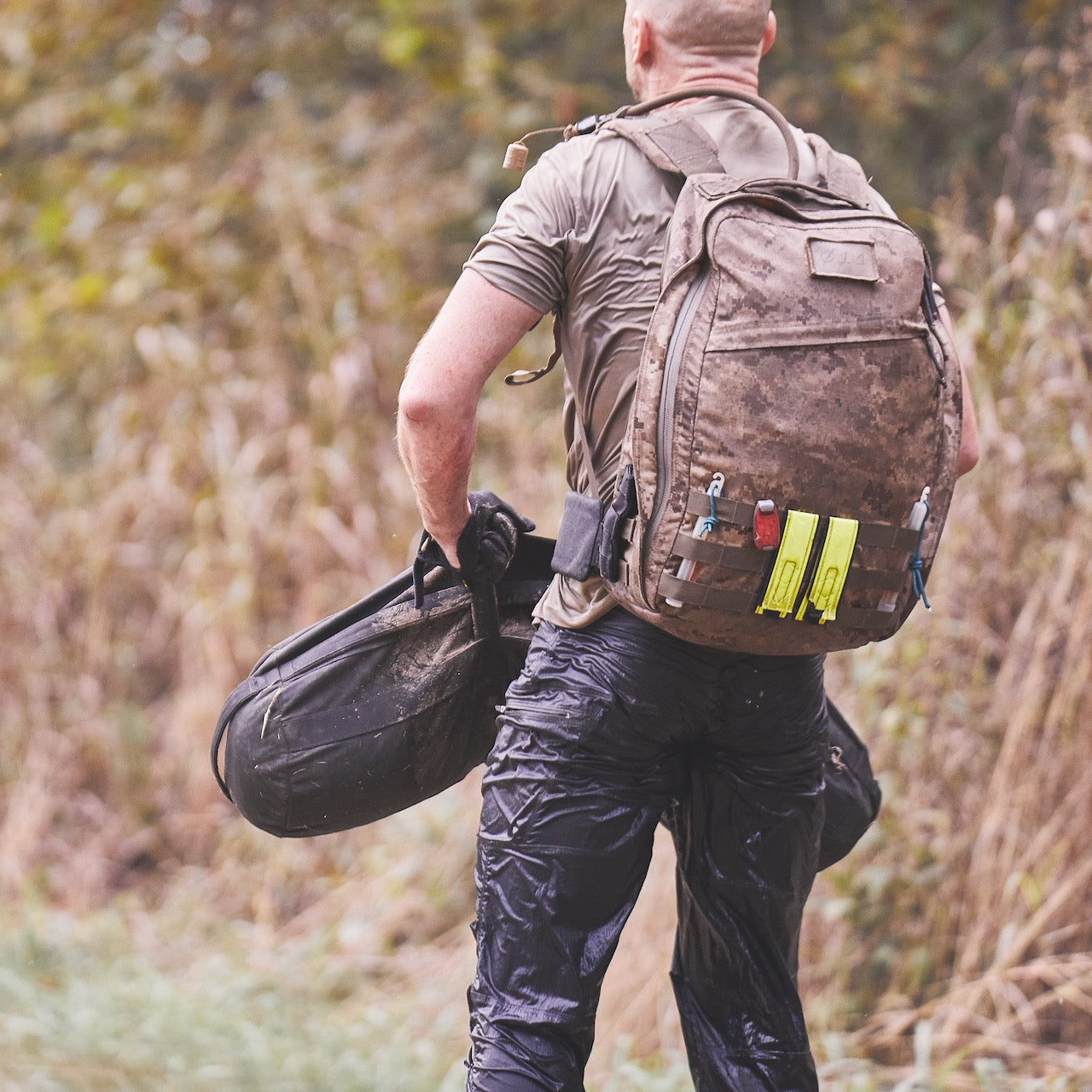 A man with wet clothes and a muddy backpack walks outdoors, carrying the Sandbag Kit (w/ Filler Bag) in one hand.