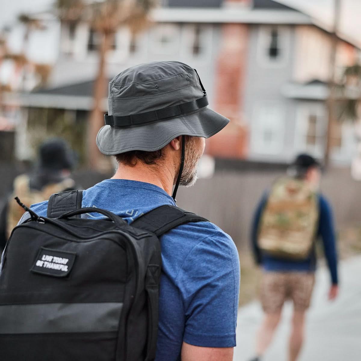 Man wearing charcoal GORUCK Boonie hat and blue shirt with black backpack on urban sidewalk