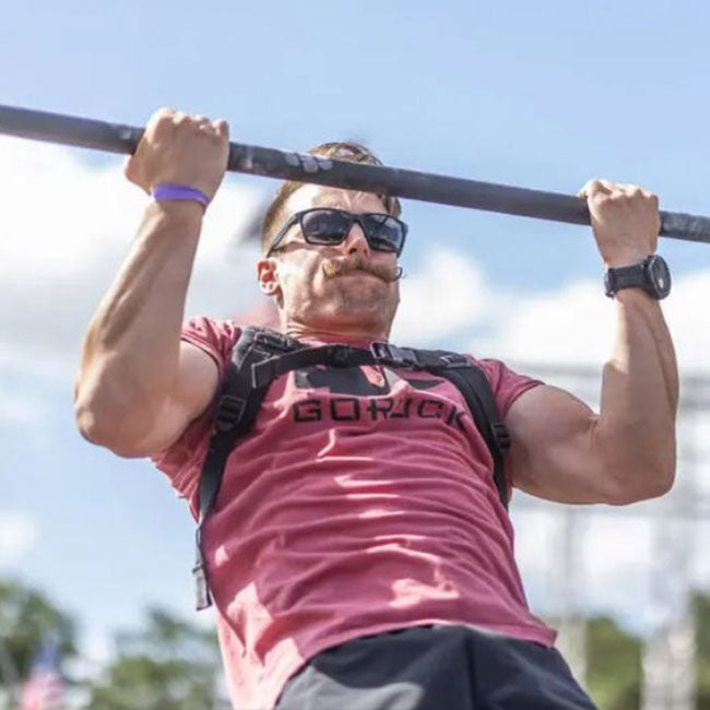Man in sunglasses and a red shirt doing a pull-up outdoors, gripping a bar with both hands.