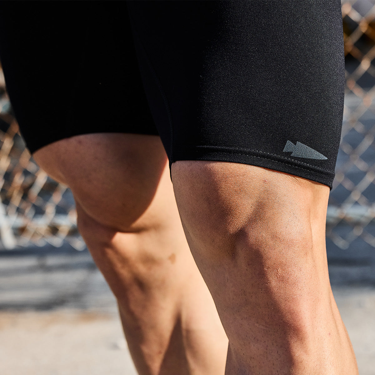 Close-up of a person's muscular thighs wearing Performance Brief black, moisture-wicking shorts, standing outdoors near a chain-link fence.