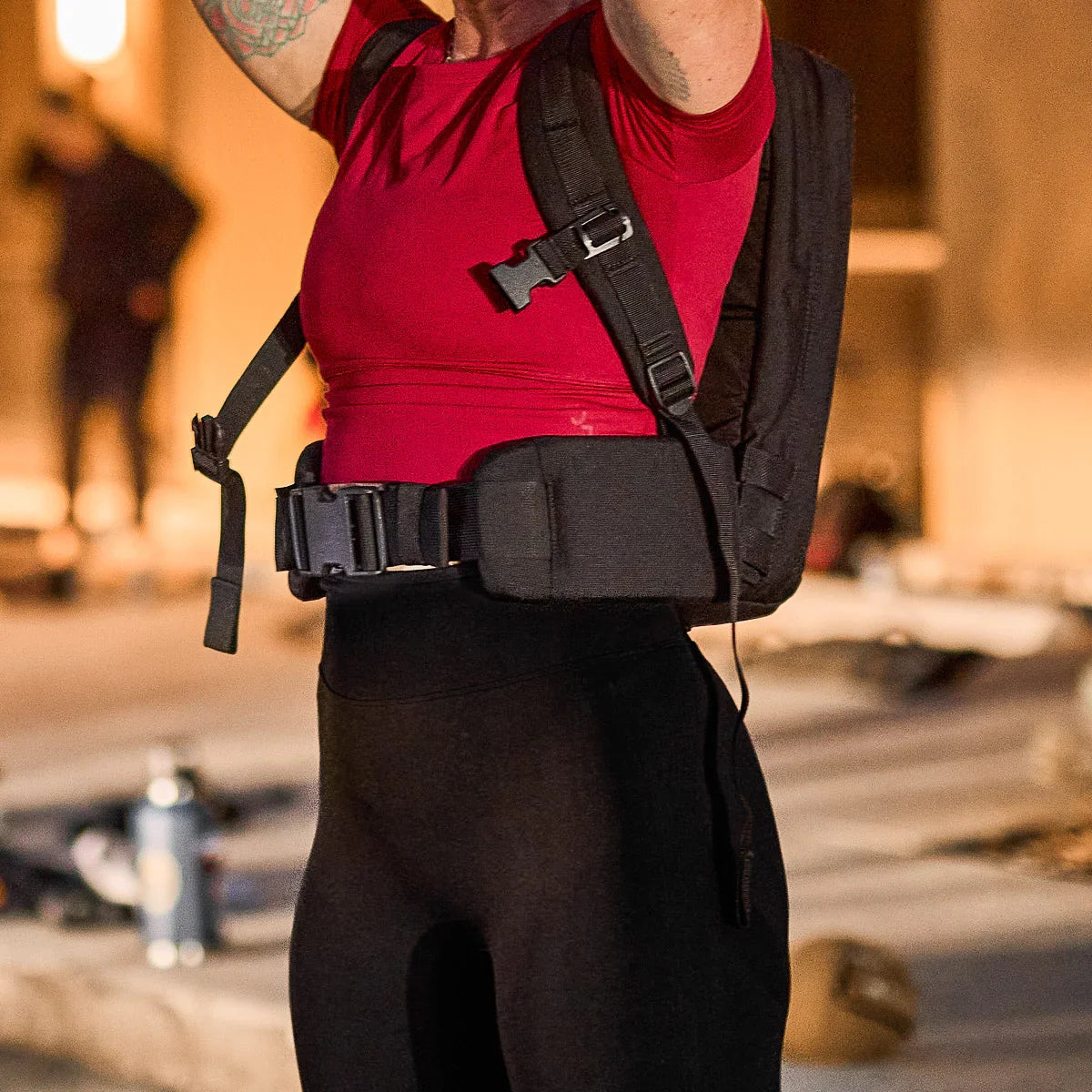 Person wearing red shirt and black GORUCK rucking pack with waist belt in outdoor urban setting