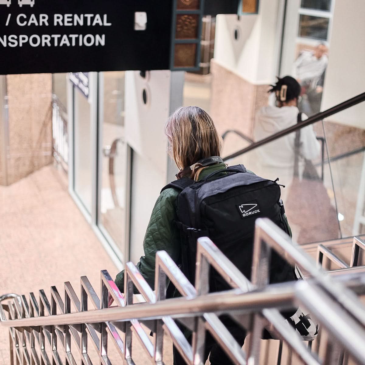 A person with the GR3 - X-PAC, a black waterproof carry-on backpack, walks down stairs in a transit area near a car rental sign.
