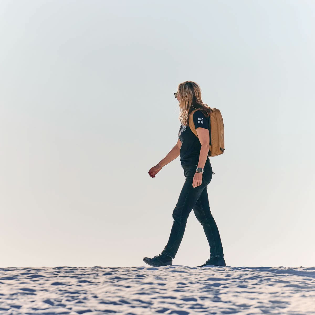 A person with long hair and a lightweight GR0 - X-PAC rucksack walks on sand under a clear sky.