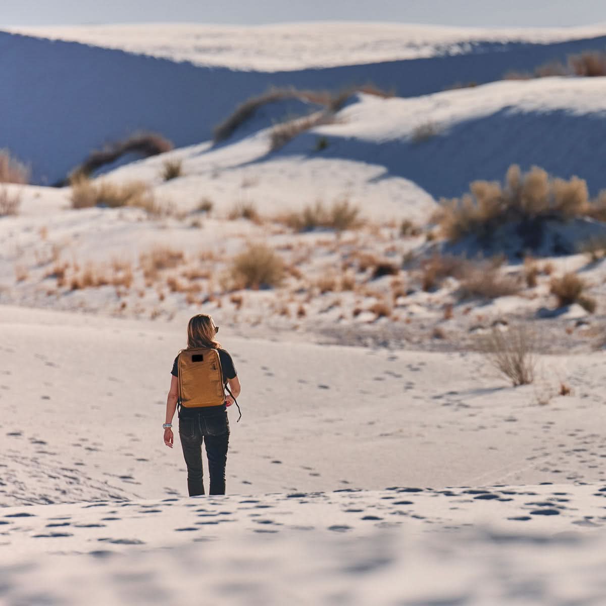 A person carrying the lightweight GR0 - X-PAC rucksack walks alone through sandy dunes with sparse bushes under a bright sky.
