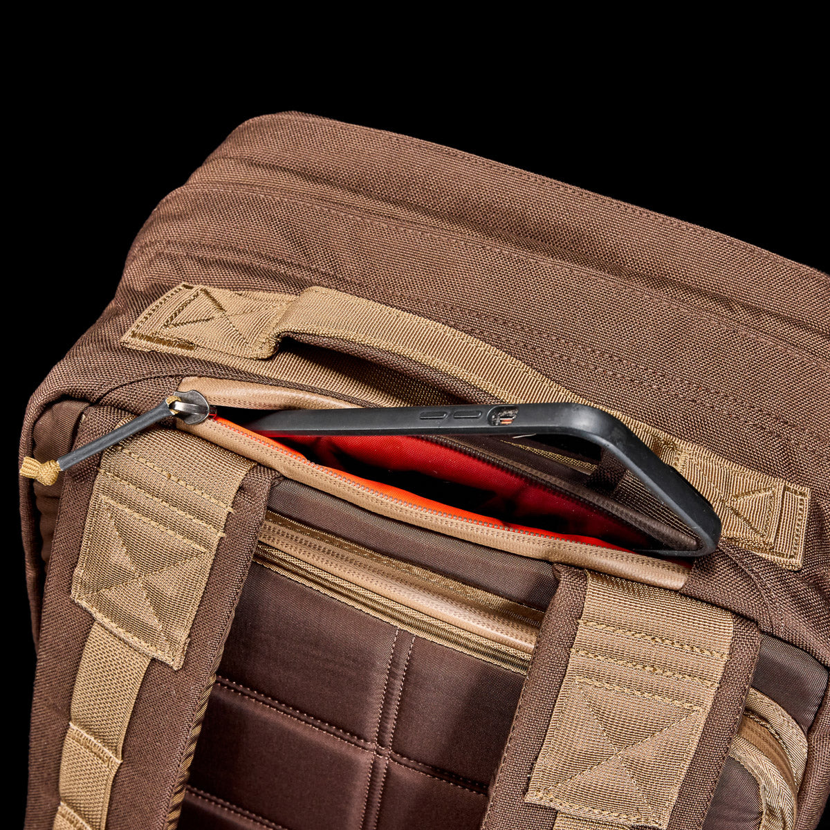 Close-up of brown GORUCK rucksack with padded straps and zippered pocket holding a smartphone with black case