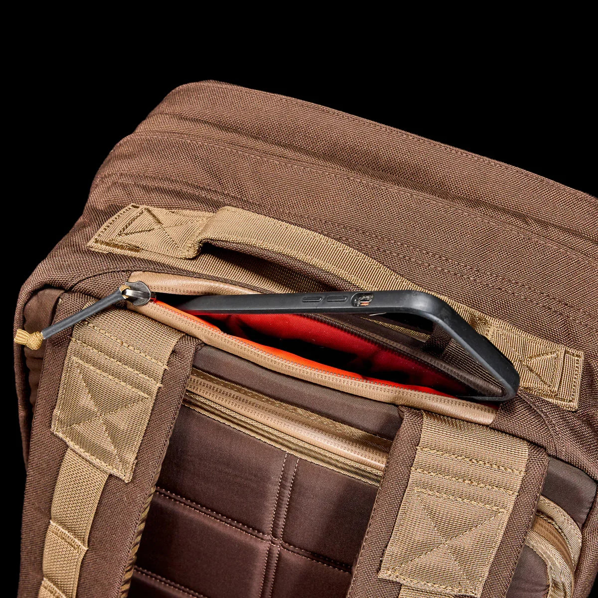 Close-up of brown GORUCK rucksack with padded straps and zippered pocket holding a smartphone with black case
