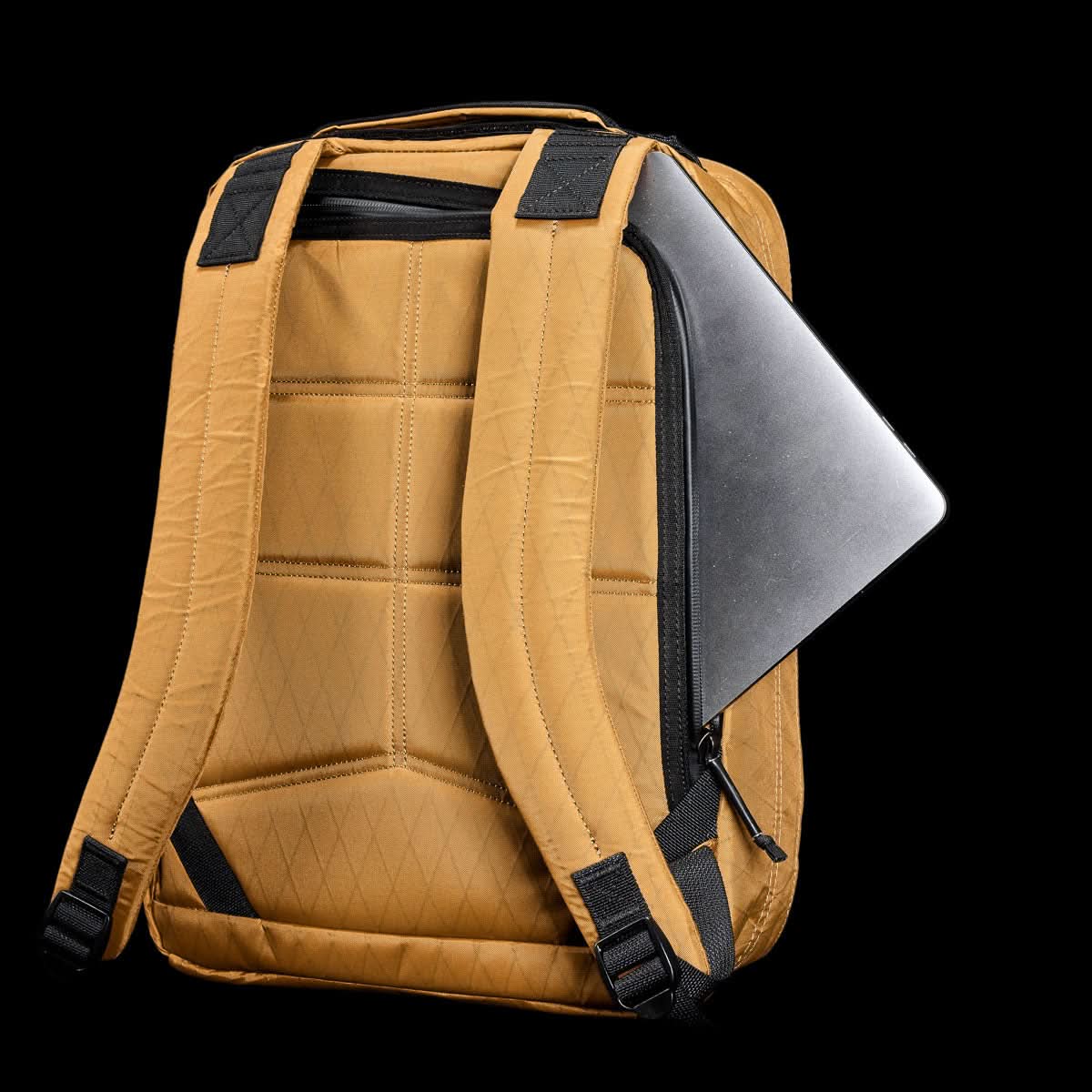 The GR0 - X-PAC is a tan, waterproof rucksack with black straps and a partially visible laptop in the side zipper pocket, shown on a black background.